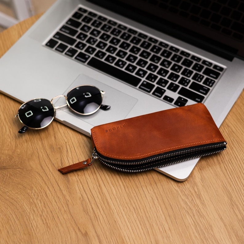 May include: A brown leather zippered case for glasses with the word "DIVORE" embossed on the front. The case is sitting on a wooden surface next to a laptop and sunglasses.