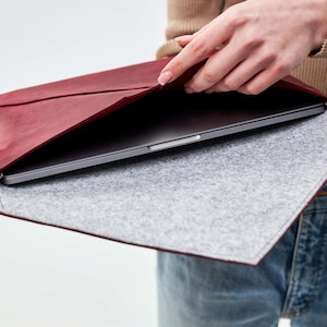 May include: A burgundy leather laptop sleeve with a gray felt lining. The sleeve is open and a laptop is inside.