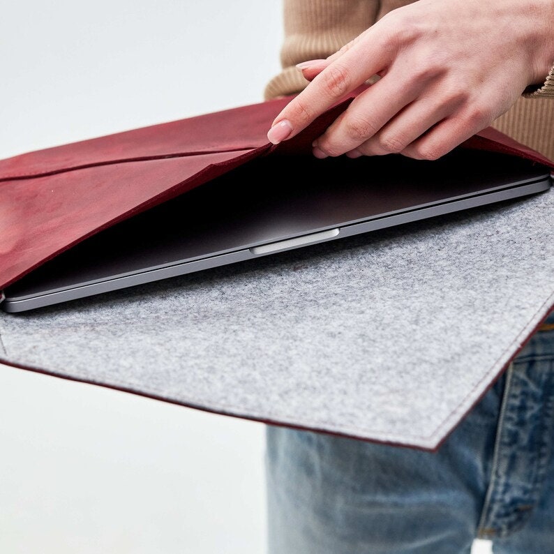 May include: A burgundy leather laptop sleeve with a gray felt lining. The sleeve is open and a laptop is inside.