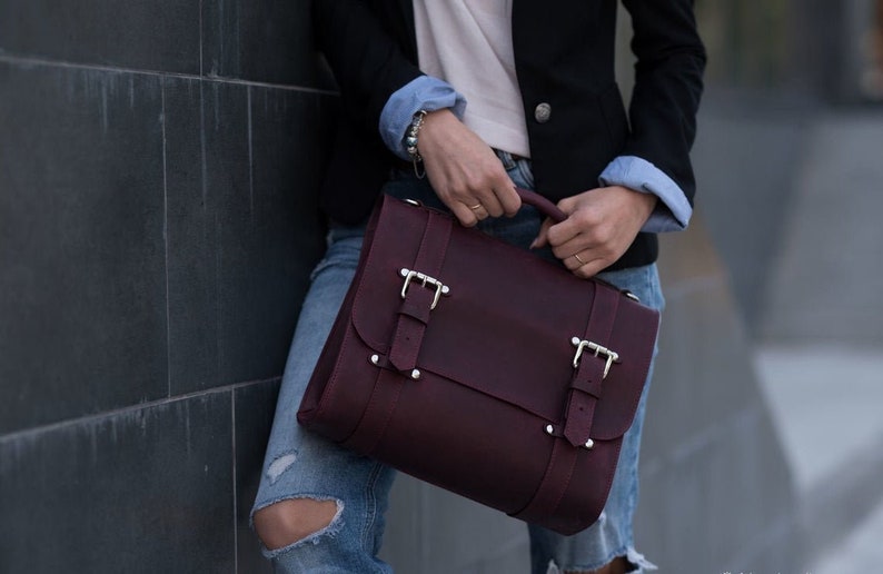 May include: A burgundy leather satchel bag with silver buckles and a flap closure. The bag is held by a person wearing a black blazer and blue jeans.