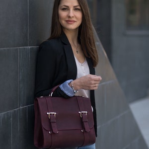 May include: A woman wearing a black blazer, a white shirt, and blue jeans is holding a burgundy leather satchel bag with silver buckles.