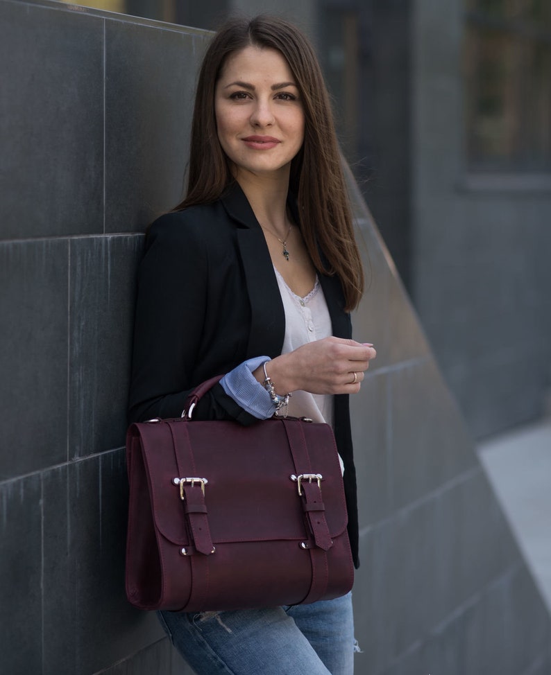 May include: A woman wearing a black blazer, a white shirt, and blue jeans is holding a burgundy leather satchel bag with silver buckles.