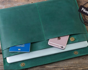 May include: A green leather laptop sleeve with a passport and a phone inside. The sleeve has a flap that closes with a snap. The sleeve is handmade and has a label that says "Pikore Workshop".