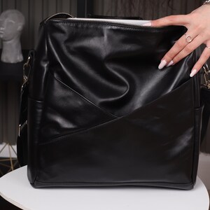 May include: A black leather satchel bag with a zipper closure and a long adjustable strap. The bag has a geometric design on the front.