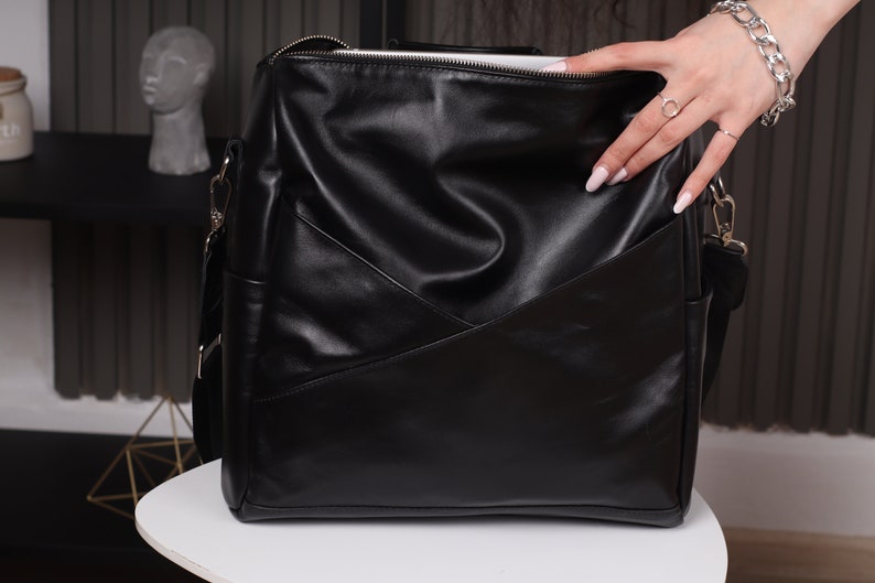May include: A black leather satchel bag with a zipper closure and a long adjustable strap. The bag has a geometric design on the front.