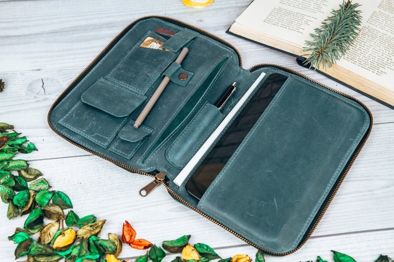 May include: A teal leather zippered portfolio with multiple pockets and slots for holding pens, cards, and a tablet. The portfolio is open and lying flat on a white wooden surface.