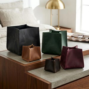Puede incluir: Una colección de cinco cajas de almacenamiento cuadradas hechas de cuero liso en varios tamaños y colores, incluyendo negro, verde oscuro, tostado, marrón oscuro y burdeos, con costuras visibles a lo largo de los bordes.