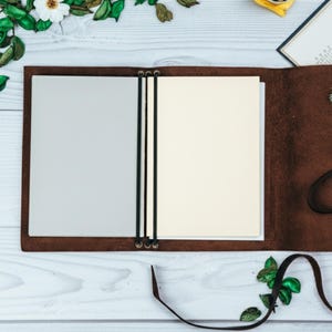 May include: Open brown leather journal with cream and gray pages. The journal is held together with black elastic bands and has a long leather strap. The journal is on a white wooden surface with decorative floral accents.