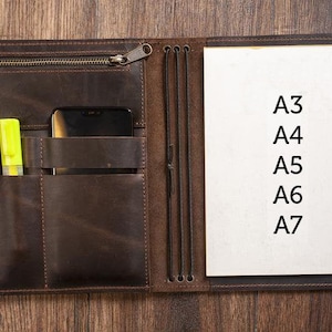 K&ouml;nnte beinhalten: Ein braunes Ledernotizbuch mit Rei&szlig;verschluss, mehreren Taschen und Gummib&auml;ndern. Das Notizbuch ist ge&ouml;ffnet und zeigt eine leere wei&szlig;e Seite mit dem Text "A3, A4, A5, A6, A7" in schwarzer Tinte.