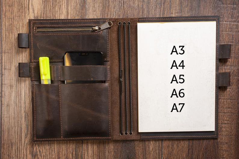 K&ouml;nnte beinhalten: Ein braunes Ledernotizbuch mit Rei&szlig;verschluss, mehreren Taschen und Gummib&auml;ndern. Das Notizbuch ist ge&ouml;ffnet und zeigt eine leere wei&szlig;e Seite mit dem Text "A3, A4, A5, A6, A7" in schwarzer Tinte.