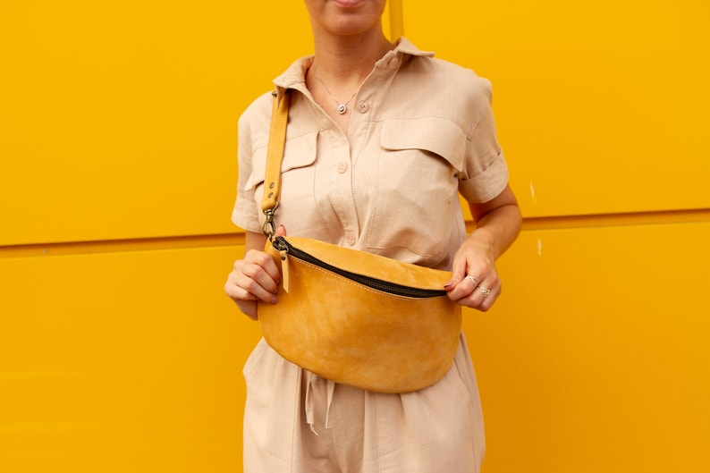 May include: Mustard yellow leather fanny pack with a black zipper and adjustable strap. The bag is worn across the body. The background is a solid yellow wall.