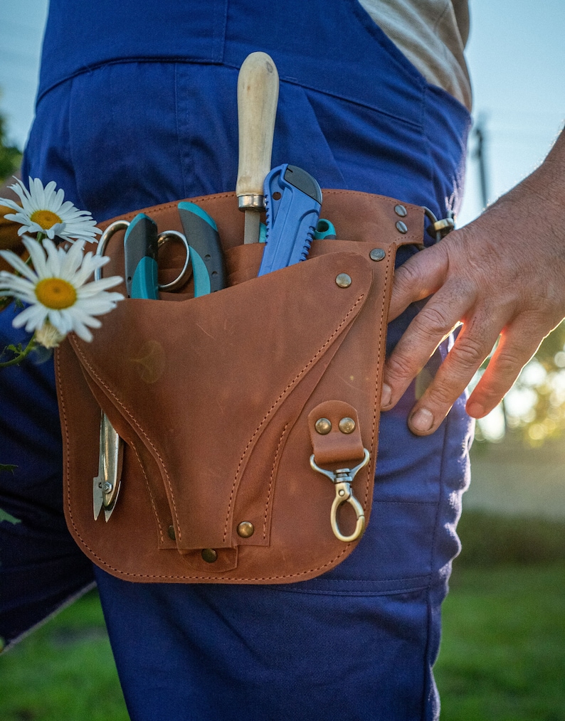May include: A brown leather tool belt with a variety of gardening tools, including a pruning shears, a knife, and a saw. The belt is worn by a person wearing blue overalls.
