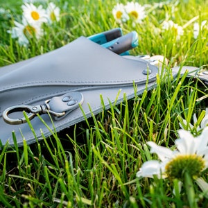 May include: A gray leather tool belt with a metal clasp and a blue and black tool hanging from it. The belt is lying on a bed of green grass with white daisies.