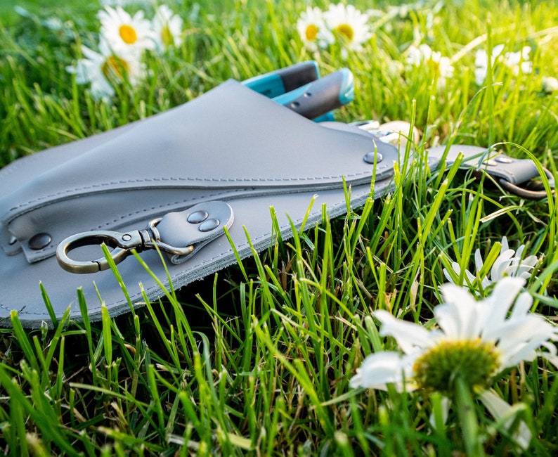 May include: A gray leather tool belt with a metal clasp and a blue and black tool hanging from it. The belt is lying on a bed of green grass with white daisies.