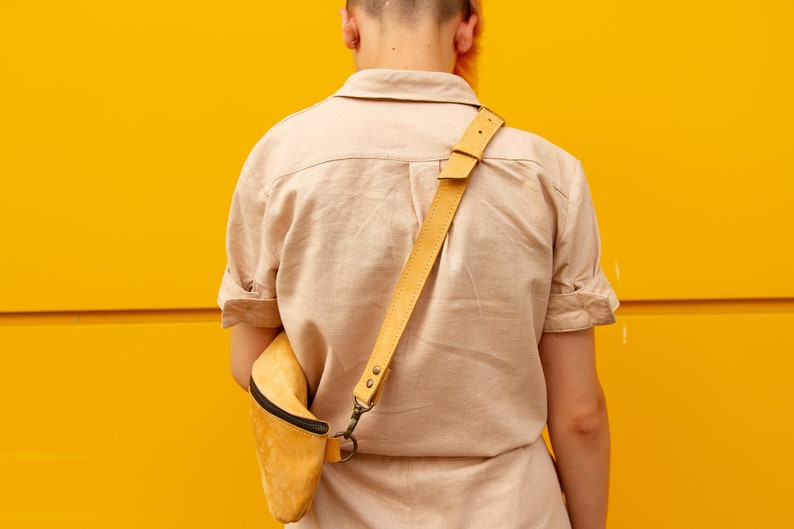 May include: A light yellow leather crossbody bag with a black zipper and a matching adjustable strap. The bag is worn over the shoulder. The background is a solid yellow color.