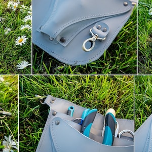 May include: A gray leather gardening tool belt with multiple pockets and a metal clip. The belt is lying on a bed of green grass with white flowers.