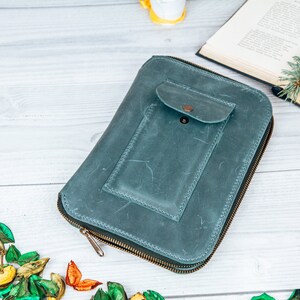 May include: A teal blue leather zippered wallet with a smaller zippered pocket on the front. The wallet is lying on a white wooden surface.