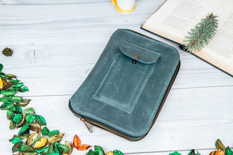 May include: A teal blue leather zippered wallet with a smaller zippered pocket on the front. The wallet is lying on a white wooden surface.
