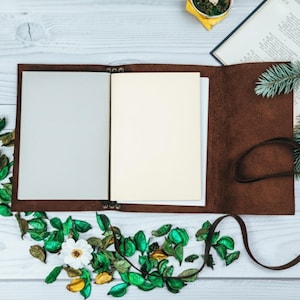 May include: An open, brown leather journal with blank pages, lying on a white wooden surface. The journal is surrounded by green and white decorative leaves and flowers. A brown leather strap is wrapped around the journal.