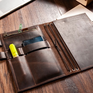 May include: A brown leather notebook with multiple pockets and elastic closures. The notebook has a zippered pocket on the front and a phone pocket on the inside. A yellow highlighter and a black phone are visible in the pockets.