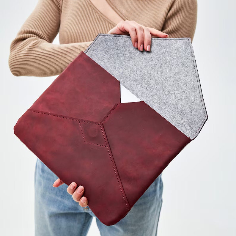 May include: A burgundy leather laptop sleeve with a gray felt lining. The sleeve is open and the felt lining is visible.