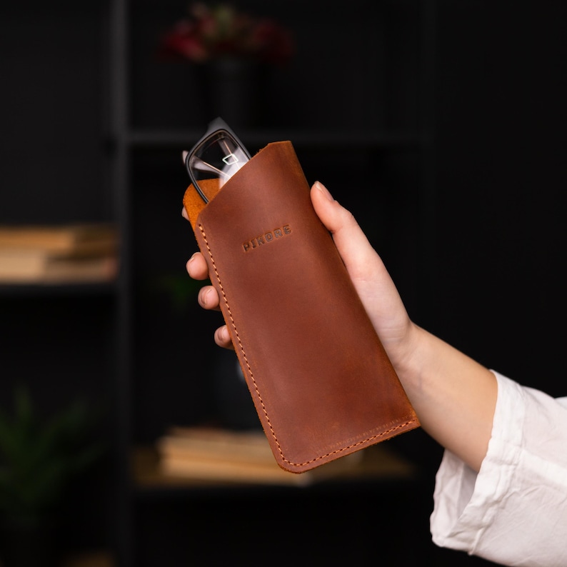 May include: A brown leather glasses case with the word "MINIMAL" embossed on the front. The case is closed and held in a hand.