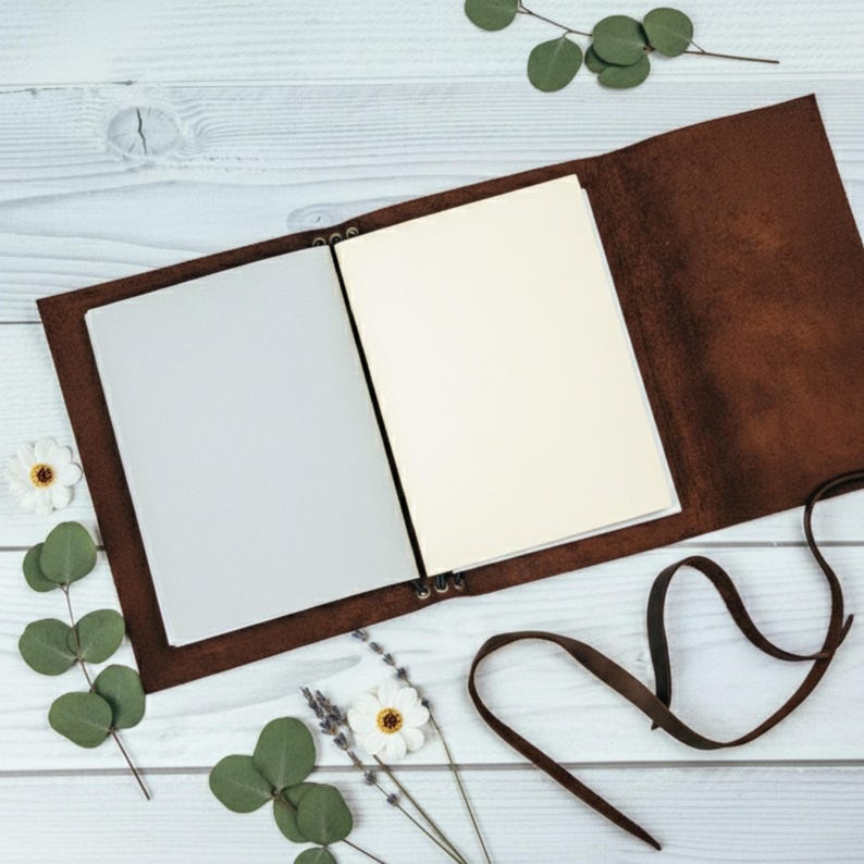 May include: Open brown leather journal with blank pages. The journal is lying open on a white wooden surface, with decorative flowers and greenery around it. A brown leather strap is visible.
