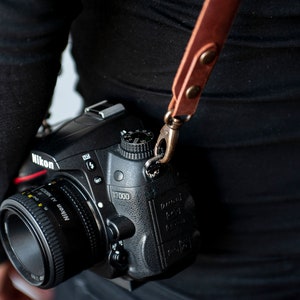 K&ouml;nnte beinhalten: Eine schwarze Nikon D7000 DSLR-Kamera mit einem schwarzen Objektiv. Die Kamera h&auml;ngt an einem braunen Lederriemen.