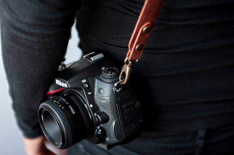 K&ouml;nnte beinhalten: Eine schwarze Nikon D7000 DSLR-Kamera mit einem schwarzen Objektiv. Die Kamera h&auml;ngt an einem braunen Lederriemen.