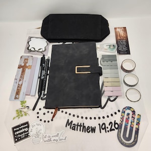 May include: A black notebook with a gold clasp, a black pencil case, two pens, a cross bookmark, a bookmark with text, a correction tape, a set of adhesive tabs, three rolls of washi tape, a large floral paperclip, and two stickers with text.