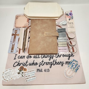 May include: A beige fabric pouch, a tan notebook, two pens, a cross bookmark, a floral bookmark, three rolls of washi tape, a set of sticky tabs, and a large pink cloth featuring the text I can do all things through Christ who strengthens me Phil. 4:13.