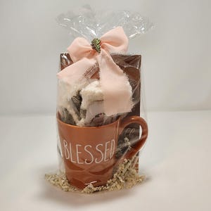May include: A burnt orange ceramic mug with the word BLESSED printed in white, packaged in clear cellophane with a light pink ribbon and a small leaf charm. Inside the mug are white fuzzy socks and a brown card, all resting on paper filler.