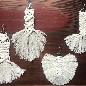 May include: Four white macrame keychains with silver key rings. The keychains are in the shape of feathers with fringe details.