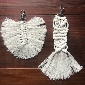 K&ouml;nnte beinhalten: Zwei wei&szlig;e Makramee-Schl&uuml;sselanh&auml;nger mit Fransen. Ein Schl&uuml;sselanh&auml;nger ist federf&ouml;rmig, der andere ist ein langes Rechteck.