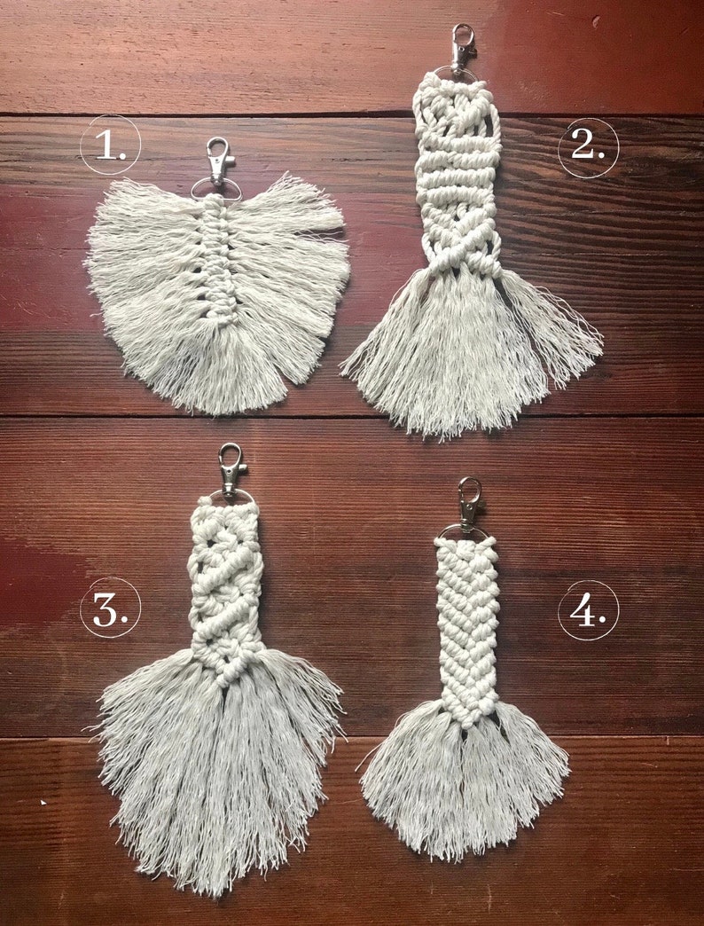 K&ouml;nnte beinhalten: Vier Makramee-Schl&uuml;sselanh&auml;nger mit Fransen, nummeriert von 1 bis 4. Die Schl&uuml;sselanh&auml;nger sind aus wei&szlig;em Kordel gefertigt und haben einen silbernen Metallring.