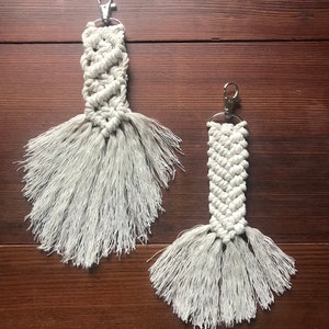 K&ouml;nnte beinhalten: Zwei wei&szlig;e Makramee-Schl&uuml;sselanh&auml;nger mit Fransen-Quasten und silbernen Schl&uuml;sselringen. Die Schl&uuml;sselanh&auml;nger h&auml;ngen an einer Holzoberfl&auml;che.