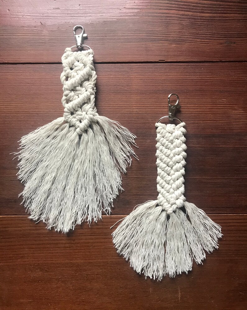 K&ouml;nnte beinhalten: Zwei wei&szlig;e Makramee-Schl&uuml;sselanh&auml;nger mit Fransen-Quasten und silbernen Schl&uuml;sselringen. Die Schl&uuml;sselanh&auml;nger h&auml;ngen an einer Holzoberfl&auml;che.
