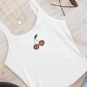 Puede incluir: Camiseta sin mangas blanca con un diseño de cerezas marrones con la inscripción "OH 10" y un tallo verde. La imagen también muestra pantalones cortos vaqueros, gafas de sol y un collar dorado.