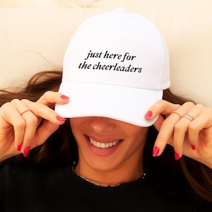 以下が含まれることがあります： 「just here for the cheerleaders」のテキストが黒い筆記体で書かれた白い野球帽。赤いマニキュアを塗った人が帽子を持っています。その人は黒いスウェットシャツを着ています。