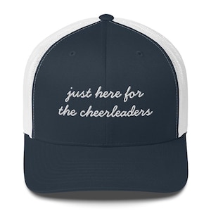 以下が含まれることがあります： 「just here for the cheerleaders」の文字が白い筆記体で刺繍された、ネイビーブルーと白のトラッカーハット。メッシュバックとカーブしたつばが特徴で、カジュアルな装いに適しています。