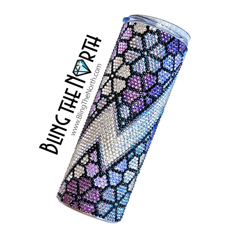 May include: A stainless steel tumbler with a purple, blue, and silver rhinestone design. The tumbler has a clear lid and is decorated with a snake skin pattern. The text "BLING THE NORTH" and "www.BlingTheNorth.com" is printed on the side of the tumbler.
