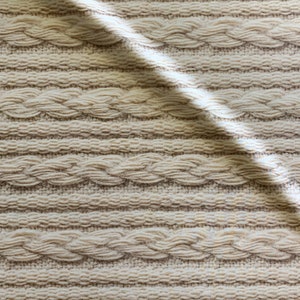 Puede incluir: Primer plano de una tela de punto de color crema con un patr&oacute;n de punto trenzado. La tela es texturizada y tiene un aspecto suave y acogedor.