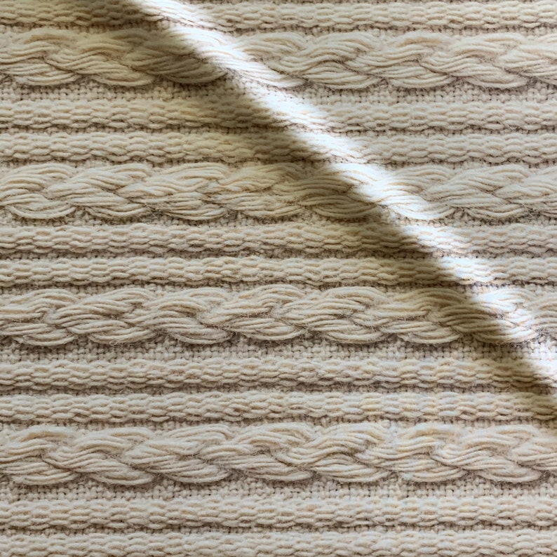 Puede incluir: Primer plano de una tela de punto de color crema con un patr&oacute;n de punto trenzado. La tela es texturizada y tiene un aspecto suave y acogedor.