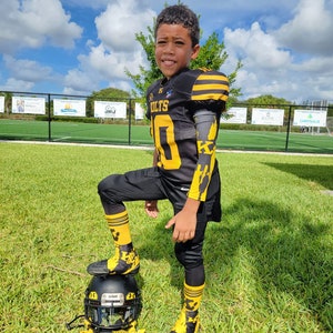 Puede incluir: Un joven jugador de f&uacute;tbol americano con un uniforme negro y dorado, que incluye casco, hombreras, camiseta con el n&uacute;mero 10 y calcetines a juego. El uniforme tiene detalles amarillos y la palabra "COLTS" es visible. Est&aacute; de pie sobre un casco en un campo de hierba.