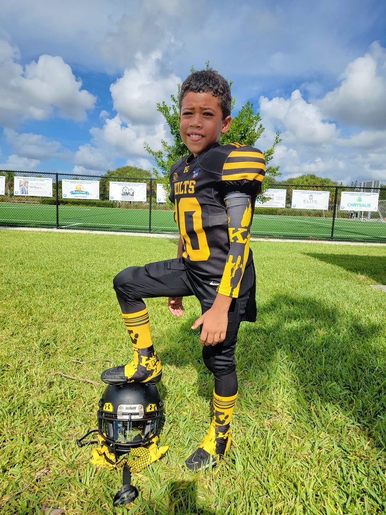 Puede incluir: Un joven jugador de f&uacute;tbol americano con un uniforme negro y dorado, que incluye casco, hombreras, camiseta con el n&uacute;mero 10 y calcetines a juego. El uniforme tiene detalles amarillos y la palabra "COLTS" es visible. Est&aacute; de pie sobre un casco en un campo de hierba.