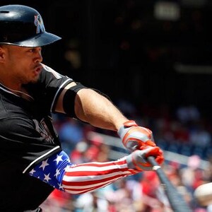 Puede incluir: Un jugador de b&eacute;isbol con uniforme negro con ribetes blancos y casco negro batea. La manga del jugador presenta un dise&ntilde;o de la bandera estadounidense. Los guantes son rojos y grises.