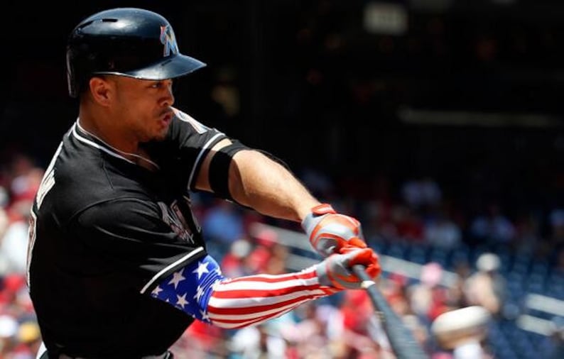 Puede incluir: Un jugador de b&eacute;isbol con uniforme negro con ribetes blancos y casco negro batea. La manga del jugador presenta un dise&ntilde;o de la bandera estadounidense. Los guantes son rojos y grises.