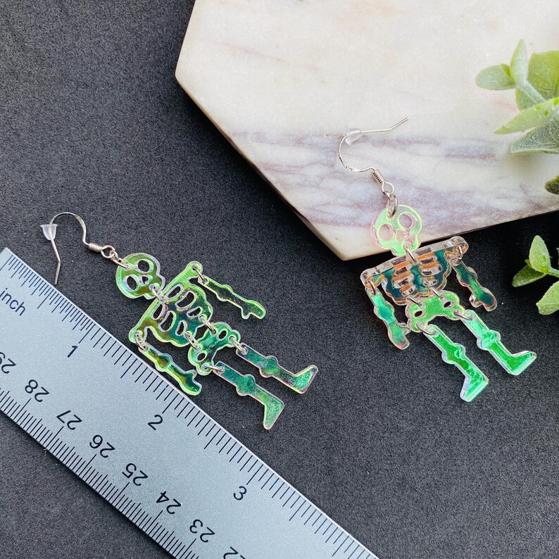 May include: A pair of iridescent skeleton earrings with articulated limbs, featuring silver-tone fishhook wires. A metal ruler is placed alongside one earring, showing the skeleton figure measures approximately 2 inches in total length.