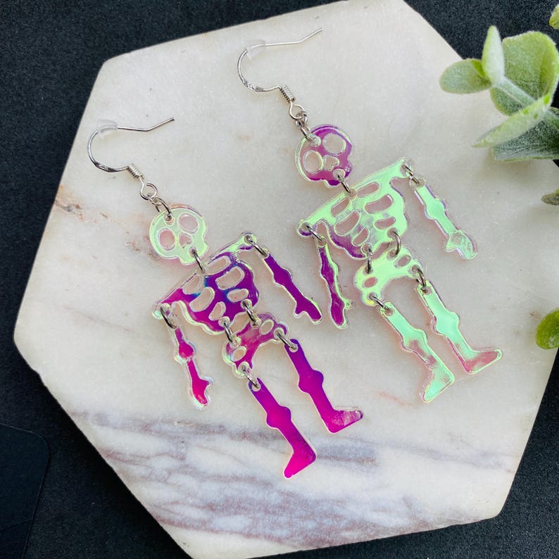 May include: A pair of dangle earrings featuring articulated skeleton figures made from iridescent, semi-transparent material. Each skeleton has a skull, ribcage, and limbs connected by silver-toned metal jump rings, suspended from silver-toned ear wires.