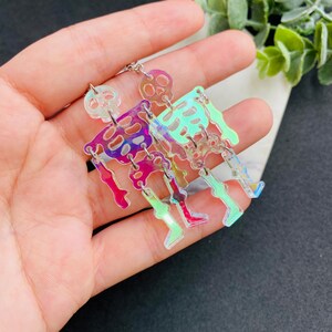 May include: A pair of dangling earrings featuring articulated skeleton figures made from iridescent, semi-transparent acrylic material. The skeletons display a shifting color spectrum of pink, purple, and green hues, connected by small silver-toned metal jump rings.